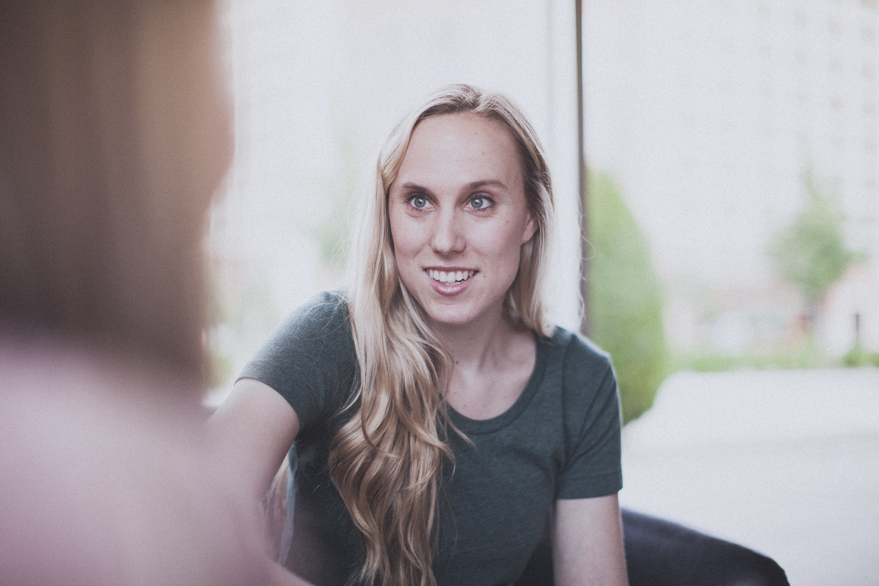 The photo features a young woman with long, wavy blonde hair. She is wearing a dark green short-sleeved shirt and is sitting indoors, seemingly engaged in conversation with someone whose blurred shoulder is visible in the foreground. The background outside the window is slightly out of focus, showing some greenery and a building, suggesting a casual and comfortable setting. The woman's expression is warm and attentive, indicating she is actively listening or engaging in a friendly conversation. The overall ambiance of the photo is bright and airy, with natural light illuminating the scene.